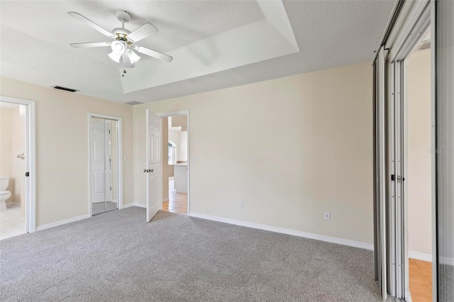 unfurnished bedroom featuring a raised ceiling, a closet, ceiling fan, connected bathroom, and light carpet