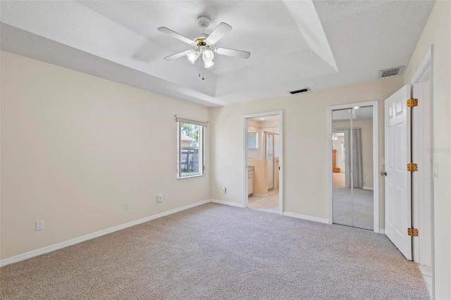 unfurnished bedroom featuring ceiling fan, light carpet, a tray ceiling, ensuite bath, and a textured ceiling