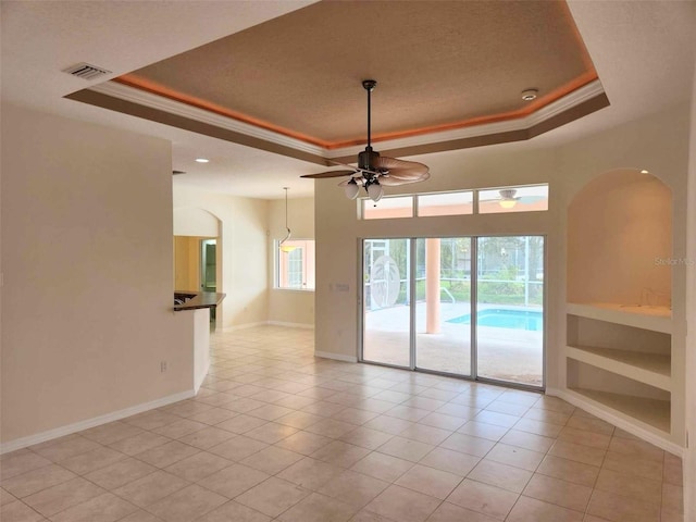 tiled empty room with a tray ceiling, built in shelves, ceiling fan, and crown molding