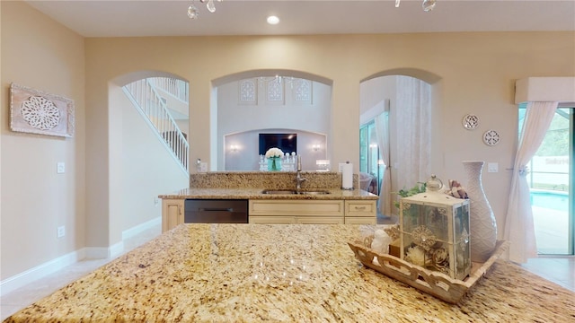 kitchen with light stone counters, cream cabinetry, a sink, dishwasher, and baseboards