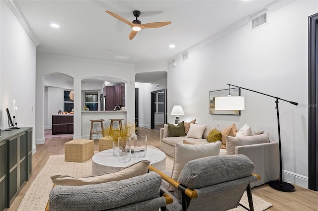 living room featuring light wood finished floors, visible vents, arched walkways, a ceiling fan, and ornamental molding