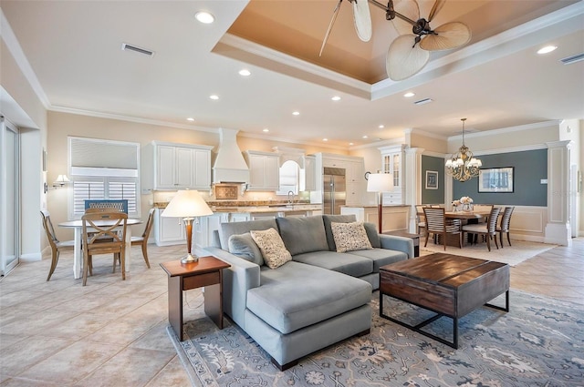 living room with ceiling fan with notable chandelier, sink, a tray ceiling, light tile patterned flooring, and ornamental molding