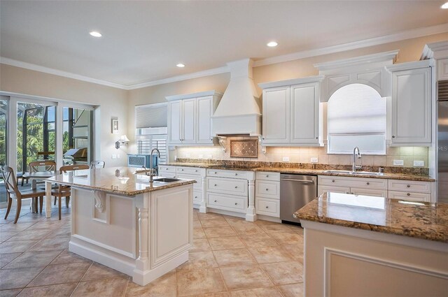 kitchen with dishwasher, premium range hood, sink, a center island with sink, and white cabinets