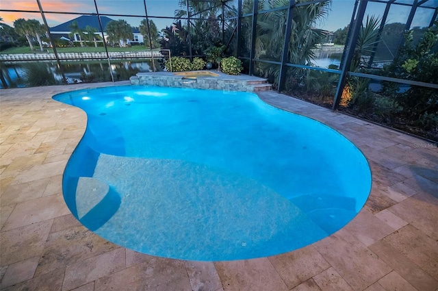 pool at dusk featuring glass enclosure, a water view, and an in ground hot tub