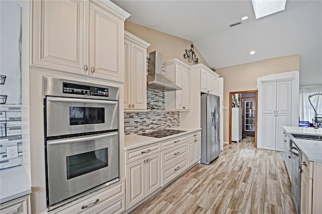 kitchen with light hardwood / wood-style flooring, lofted ceiling with skylight, stainless steel appliances, wall chimney range hood, and decorative backsplash