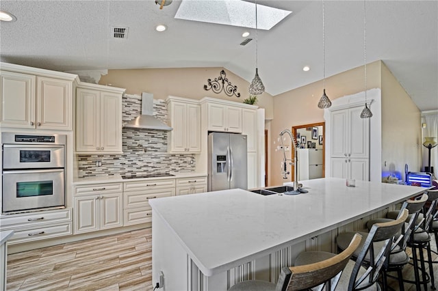 kitchen with a kitchen island with sink, a kitchen bar, stainless steel appliances, wall chimney exhaust hood, and light hardwood / wood-style floors