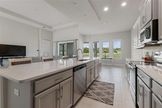 kitchen featuring gray cabinets, light stone counters, sink, appliances with stainless steel finishes, and a center island with sink