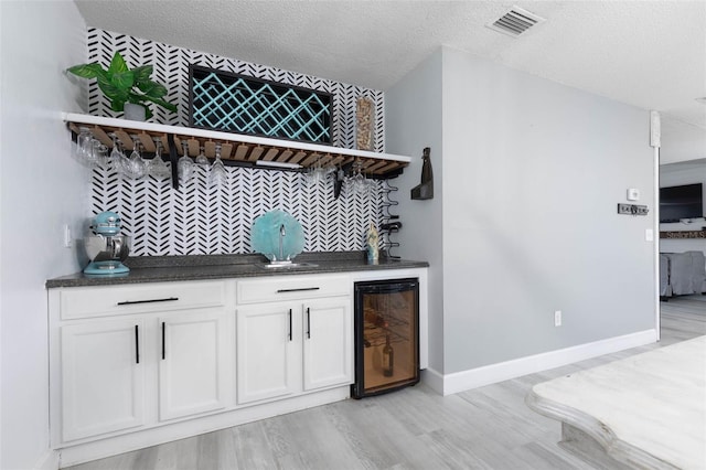 bar featuring light wood-type flooring, wine cooler, a textured ceiling, white cabinets, and backsplash