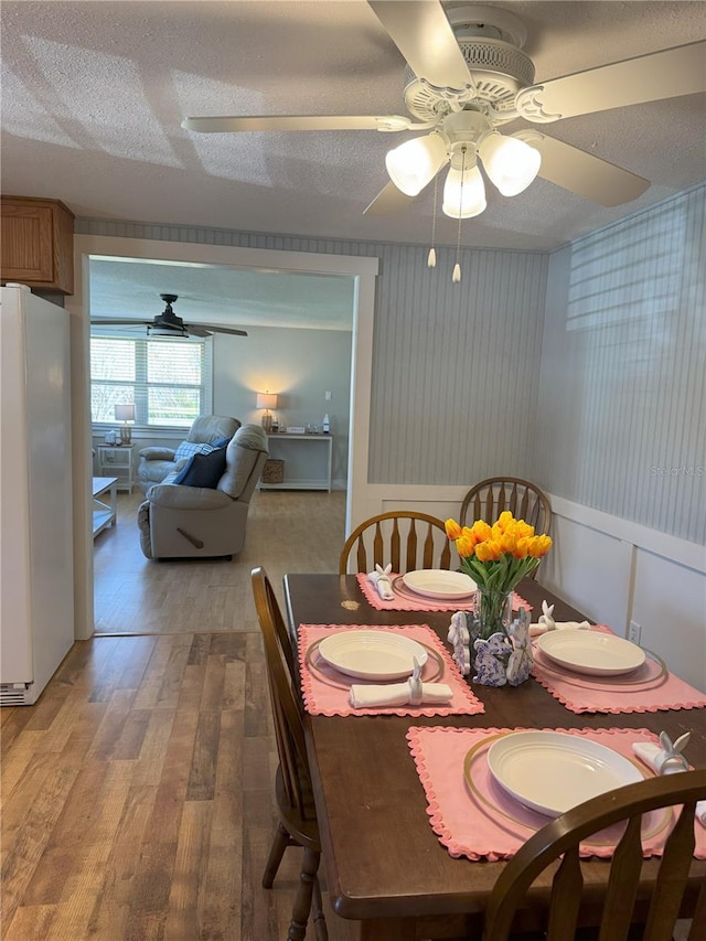 dining area featuring a textured ceiling, a ceiling fan, light wood-type flooring, wainscoting, and wallpapered walls