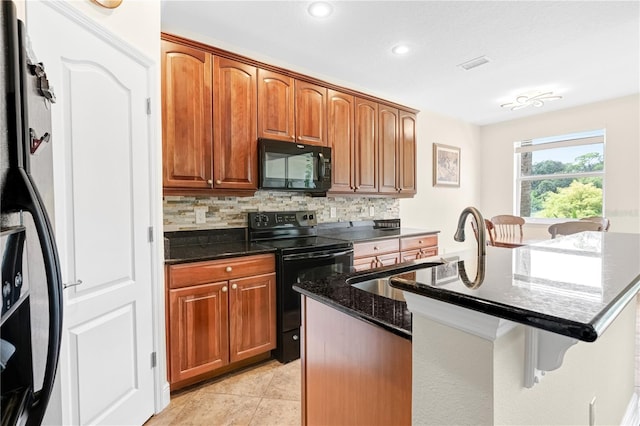 kitchen featuring sink, black appliances, a center island with sink, backsplash, and dark stone countertops