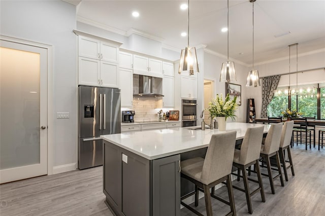 kitchen with a large island with sink, appliances with stainless steel finishes, wall chimney range hood, white cabinetry, and decorative light fixtures