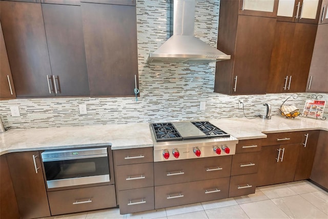 kitchen with wall chimney exhaust hood, light tile patterned floors, tasteful backsplash, light stone counters, and stainless steel gas cooktop