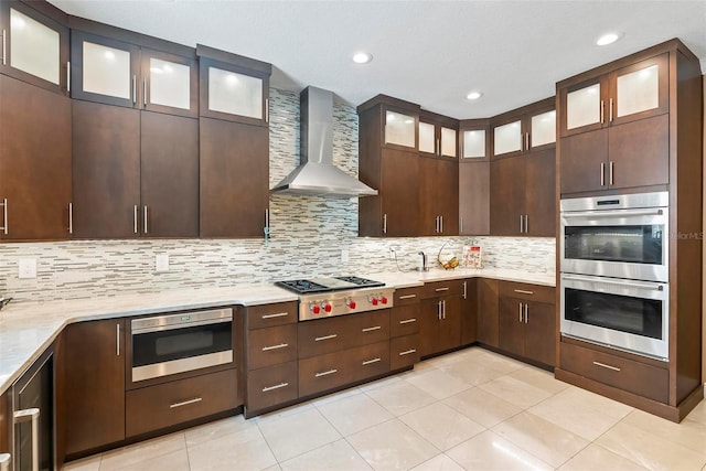 kitchen with backsplash, wall chimney range hood, light tile patterned floors, appliances with stainless steel finishes, and dark brown cabinetry