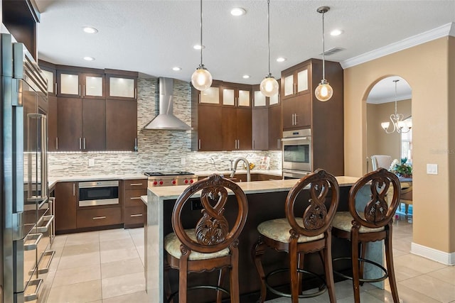 kitchen with dark brown cabinets, stainless steel appliances, wall chimney range hood, light tile patterned floors, and an island with sink