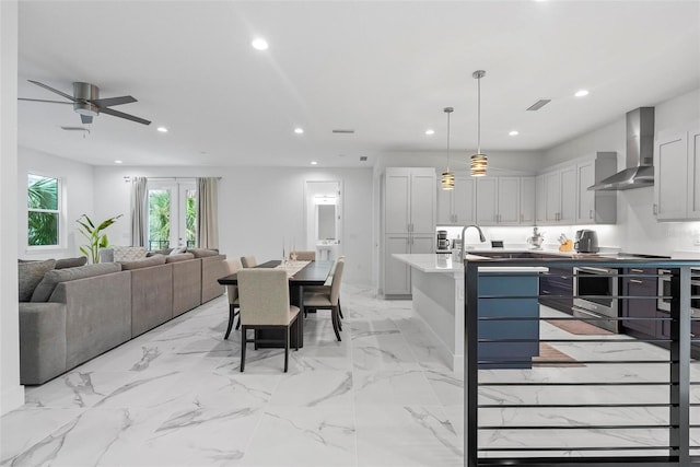 kitchen with an island with sink, white cabinets, ceiling fan, decorative light fixtures, and wall chimney range hood