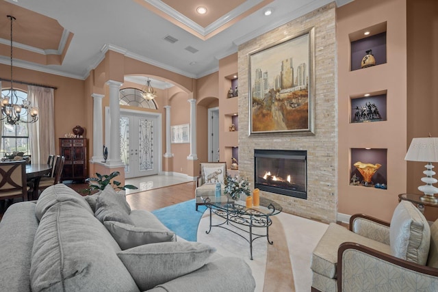 living room with light wood-type flooring, decorative columns, a tray ceiling, crown molding, and a stone fireplace