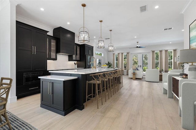 kitchen featuring a large island with sink, wall chimney range hood, light hardwood / wood-style floors, hanging light fixtures, and a breakfast bar area