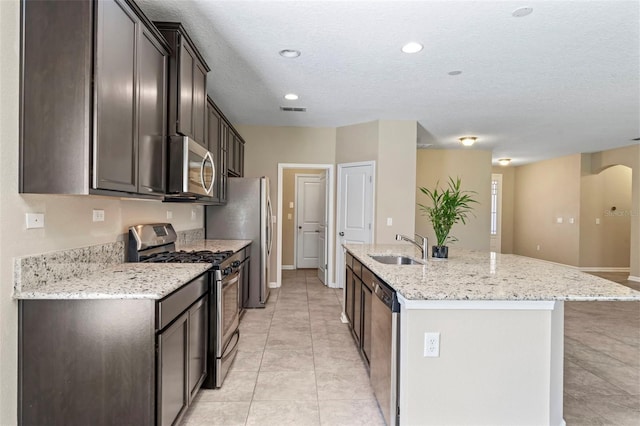 kitchen featuring sink, light tile patterned floors, stainless steel appliances, light stone counters, and an island with sink