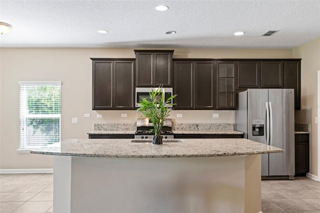 kitchen with light stone counters, a kitchen island with sink, stainless steel appliances, a sink, and dark brown cabinets