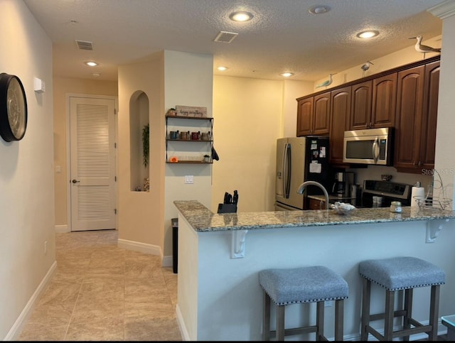 kitchen featuring a kitchen bar, light stone counters, a textured ceiling, kitchen peninsula, and stainless steel appliances