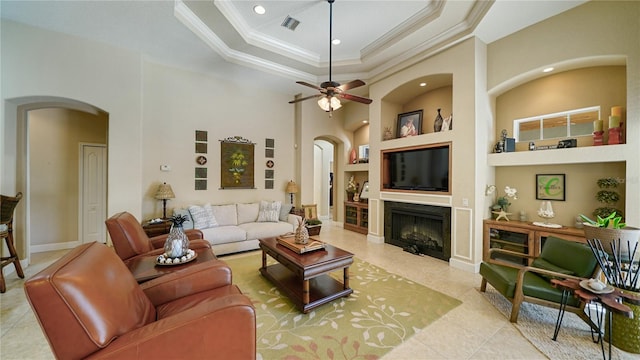 tiled living room featuring a raised ceiling, ceiling fan, a high ceiling, built in features, and crown molding