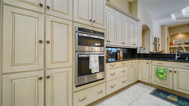 kitchen with stainless steel double oven, cream cabinetry, dark stone countertops, sink, and light tile patterned floors