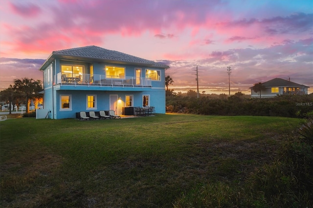 back of house at dusk featuring a balcony, stucco siding, and a yard