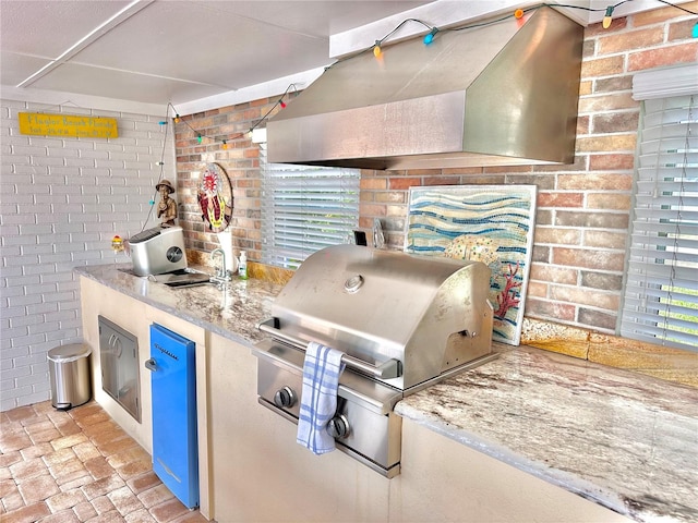 kitchen with ventilation hood, sink, stainless steel refrigerator, light stone counters, and brick wall