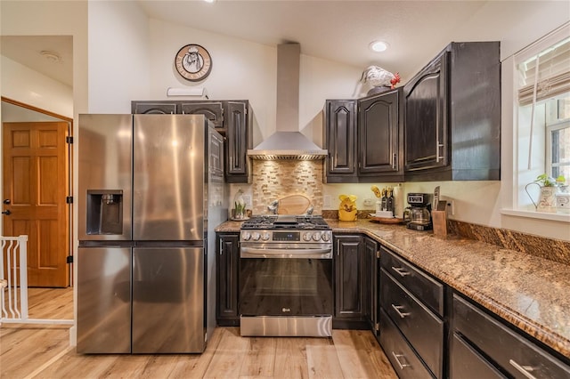 kitchen with stainless steel appliances, vaulted ceiling, wall chimney exhaust hood, light wood-type flooring, and dark stone countertops