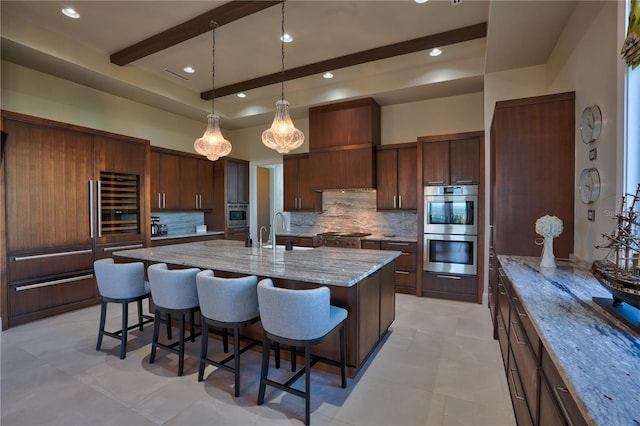 kitchen featuring stainless steel gas stovetop, a large island with sink, multiple ovens, light stone countertops, and a breakfast bar area