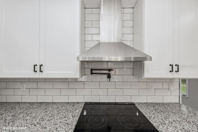 kitchen featuring black electric stovetop, wall chimney exhaust hood, white cabinetry, and light stone countertops