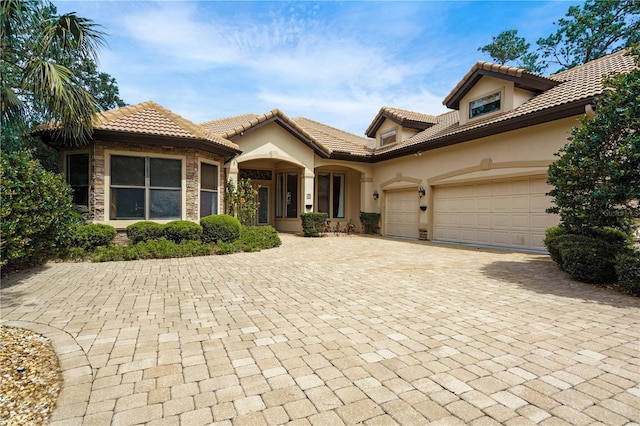 mediterranean / spanish home featuring decorative driveway, a tile roof, stucco siding, an attached garage, and stone siding