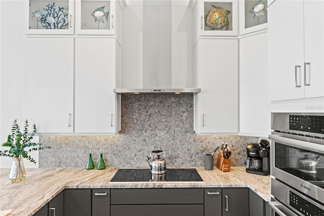 kitchen featuring decorative backsplash, light stone countertops, black electric cooktop, wall chimney range hood, and white cabinetry