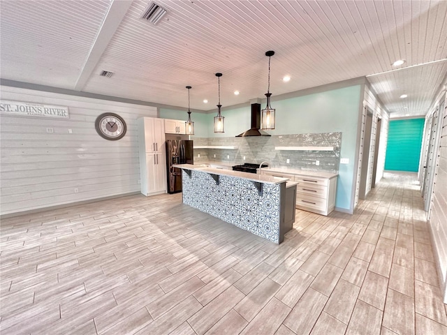 kitchen featuring light wood-type flooring, wall chimney range hood, decorative light fixtures, a center island with sink, and fridge with ice dispenser