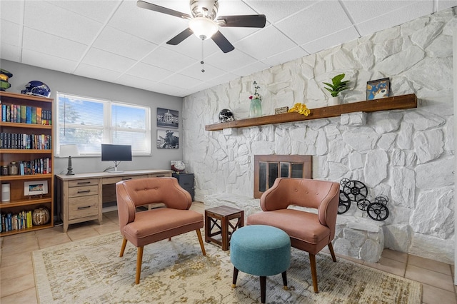 living area featuring light tile patterned floors, a paneled ceiling, a stone fireplace, and ceiling fan