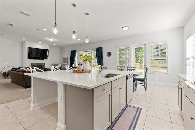 kitchen featuring dishwasher, a tile fireplace, a center island with sink, sink, and decorative light fixtures