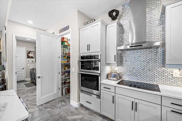 kitchen with backsplash, black electric stovetop, wall chimney exhaust hood, a textured ceiling, and double oven