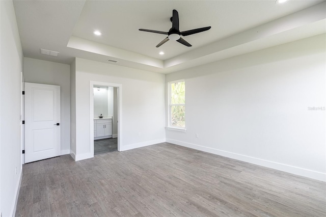 unfurnished bedroom featuring a raised ceiling, ensuite bathroom, light hardwood / wood-style flooring, and ceiling fan