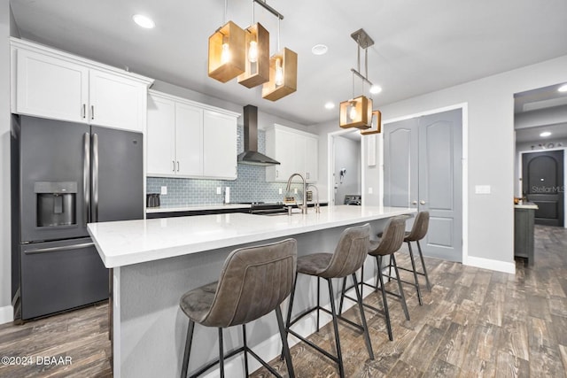 kitchen with a center island with sink, hanging light fixtures, wall chimney exhaust hood, stainless steel fridge, and white cabinetry