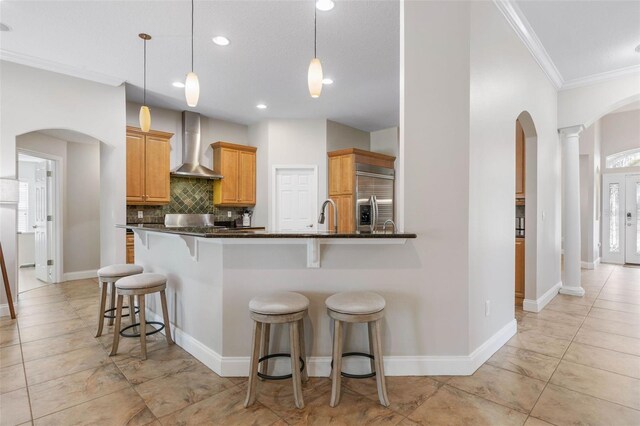 kitchen with wall chimney range hood, built in fridge, dark stone counters, decorative light fixtures, and ornamental molding