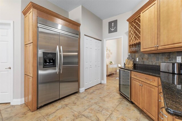 kitchen featuring appliances with stainless steel finishes, backsplash, a textured ceiling, beverage cooler, and dark stone countertops