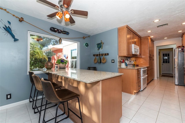 kitchen with kitchen peninsula, a kitchen bar, light stone countertops, white appliances, and ceiling fan