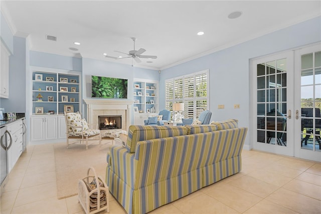 tiled living room with built in shelves, ceiling fan, french doors, a tile fireplace, and crown molding