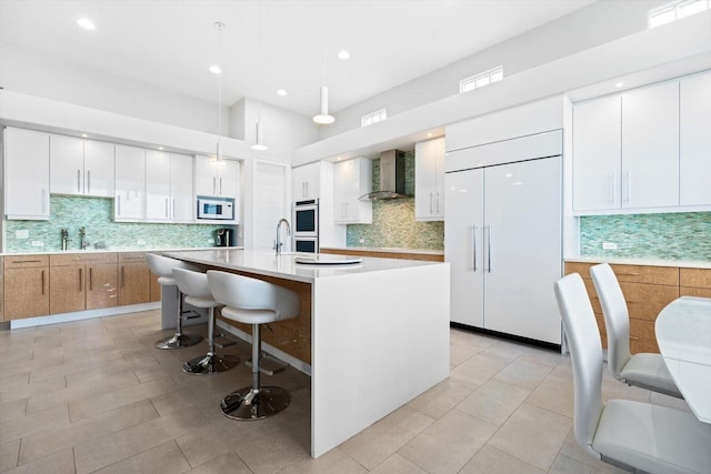 kitchen with wall chimney range hood, decorative light fixtures, paneled built in fridge, a center island with sink, and white cabinets