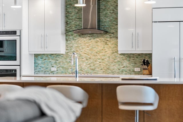 kitchen featuring double oven, white cabinets, decorative backsplash, and wall chimney exhaust hood