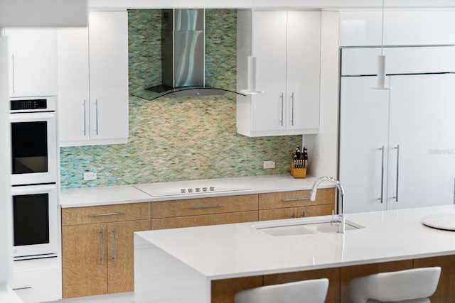 kitchen featuring tasteful backsplash, wall chimney exhaust hood, cooktop, sink, and white cabinetry