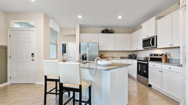 kitchen with light stone counters, stainless steel appliances, light tile patterned floors, white cabinets, and an island with sink