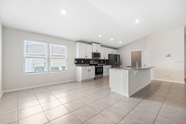 kitchen with appliances with stainless steel finishes, light stone counters, vaulted ceiling, white cabinets, and an island with sink