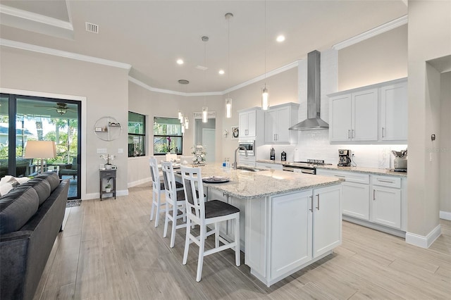 kitchen with white cabinets, a center island with sink, wall chimney range hood, and hanging light fixtures