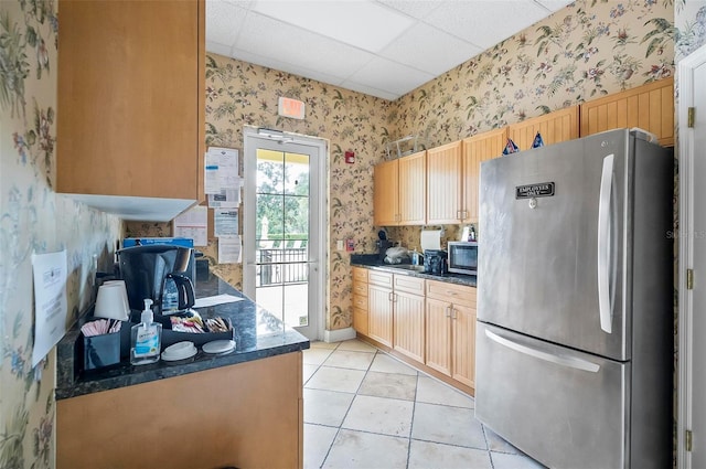 kitchen with a drop ceiling, light brown cabinets, dark stone countertops, light tile patterned flooring, and stainless steel appliances